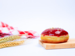 Delicious doughnut topped with red glaze and sprinkles, placed on a wooden board with wheat and red checkered cloth in background on white background