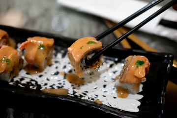 A sushi roll, possibly flambeado or a Rainbow Uramaki Roll, is held by chopsticks. The roll is...