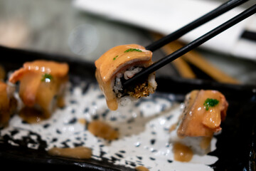 A sushi roll, possibly flambeado or a Rainbow Uramaki Roll, is held by chopsticks. The roll is...