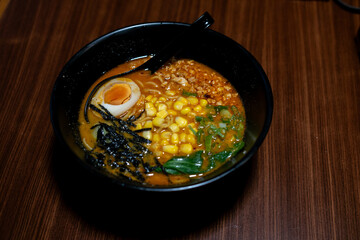 A bowl of miso ramen, possibly corn butter ramen, featuring a halved boiled egg, corn, and seaweed, presented in a black bowl on a wooden table.