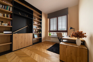 Comfortable modern home office interior featuring a wooden bookshelf filled with books, a sleek desk, and chair by the window. The room has beautiful herringbone parquet flooring and warm lighting.