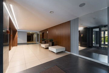 Modern residential lobby: wood panels, large mirror, tiled floor. Spacious hallway with ottomans, potted plants, and contemporary lighting. Clean, inviting interior design for an apartment building.