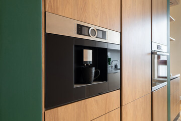 Built-in coffee machine integrated in wooden kitchen cabinets. Modern home appliance with sleek design and digital display, enhancing a contemporary residential kitchen. Integrated home technology.