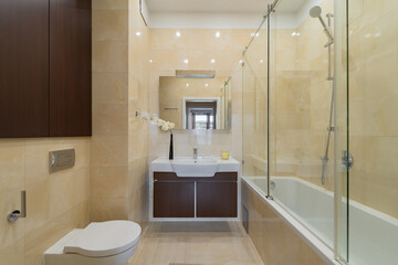 Modern home bathroom with light tiled walls and floor. Includes bathtub with glass shower screen, wall-mounted toilet, and wooden vanity. A clean, elegant residential washroom interior design.