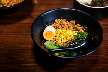 A vibrant bowl of dry ramen (mazesoba), a popular noodle dish, topped with minced meat, corn, green onions, seaweed, and a perfectly halved egg, on a wooden table.