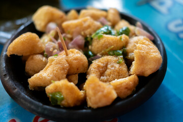 a serving of Tahu Gejrot, a popular Indonesian street food,  the fried tofu pieces mixed with a dark, watery sauce, slices of shallots, and green chili peppers in a dark bowl