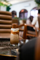 a layered iced coffee drink, likely a spiced cookie latte, served in a tall glass. It features layers of coffee, milk, and whipped topping, garnished with a spiced cookie.
