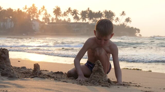 Kid playing in wet sand on a sunny beach. Boy engaged in building sandcastle on sunny beach at sunset. Use hands to mold sand and create shapes. Waves wash over sandy beach with palm trees