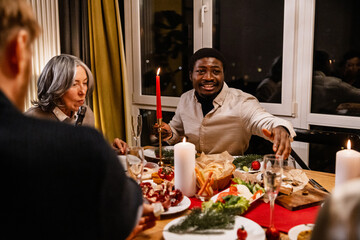 A Black adult man and a white senior woman smile and chat at a nighttime holiday dinner table, surrounded by festive food, candles, and drinks.