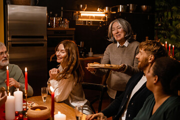 A caucasian woman serves bread to a group of White adults seated at a festive holiday dinner table with candles and champagne in a warmly lit modern kitchen.