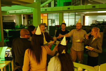 Three White men, a Black man, a white-skinned woman, and two women in birthday hats are holding their glasses of champagne together while a White woman is drinking champagne and looking at them