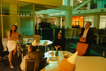 A group of diverse adults, including men and women, celebrate in an office at night wearing party hats. They sit around a table with cake and drinks, and a laptop shows a remote participant.