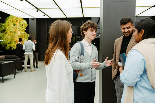 A multinational group of young people in business attire is standing indoors in a modern office lobby during an internship program, with a young White man holding notebooks and gesturing.