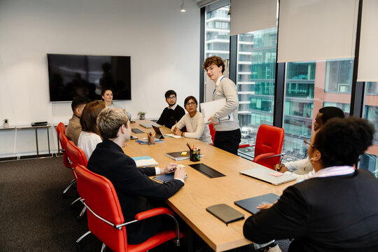 A diverse group of adults and one White teenage boy gather around a conference table in a modern office. The teen stands and speaks, holding a folder during a professional meeting.