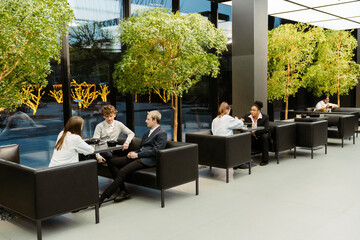 Two adult White men are sitting on a couch at a table with an adult woman, while two women are eating together at a table behind them, and a brown-skinned woman is sitting in the distance near trees