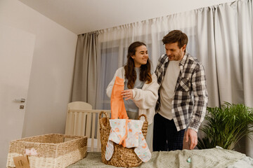 A white adult couple stands in a cozy bedroom, organizing baby clothes into a wicker basket. A crib is visible in the background, meaning they prepare for a newborn in a softly lit indoor setting.