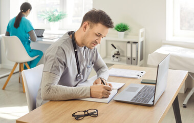 Young confident male doctor working at the desk with laptop on workplace doing paperwork in medical...
