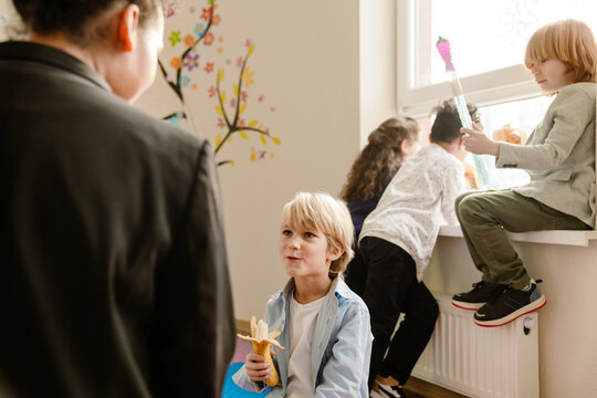 A group of school-aged children play in a classroom, one eating a banana while others explore and chat near the window, under a teacher's watchful eye. The concept of kids' interaction at school.