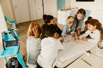 A group of school-aged children of diverse ethnicities collaborate on a large drawing with markers indoors in a classroom, participating in a creative school project.