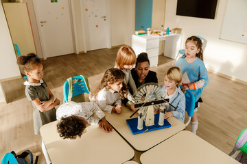 A Black woman teaches diverse school-aged children using a mechanical science device in a classroom with desks, chairs, and learning supplies in daylight.