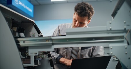 Technician Servicing ATM Machine in Modern Airport Terminal. Male Worker in Uniform Using Laptop,...
