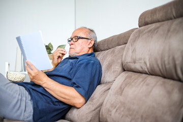 Side View Of Retired Elderly Male Sit And Leaning On Sofa Reading Book While Eating Popcorn