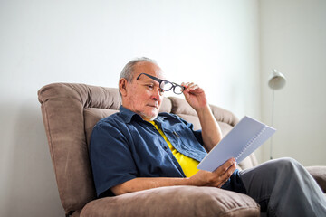 Fototapeta premium Portrait Of Retired Elderly Man Reading Book With Eye Glasses While Sitting On Sofa