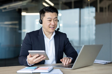 Fototapeta premium Asian businessman in a suit works remotely during a video call, using a laptop and tablet.