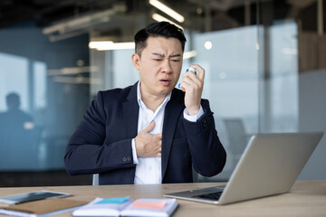 An asian businessman in a suit struggles with asthma while using an inhaler at his office desk.