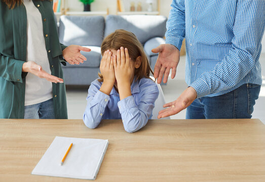 Upset child sitting at table with notebook, covering face with hands while his parents arguing or scolding in background. Father and mother criticizing kid because of educational or school problems.