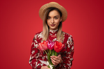 Pretty young female in stylish outfit and hat holding bouquet of tulips and looking at camera while standing on red background