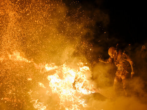 A Krampus figure stands amidst a fiery display, sparks flying in the dark. The figure is dressed in a traditional costume with a grotesque mask. Krampus and St Nicholas, 5 December in Tarvisio, Italy