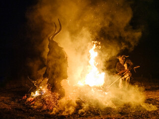 Two Krampus figures stand on either side of a bonfire, smoke and sparks fill the air. Krampus and St Nicholas, 5 December in Tarvisio, Friuli Venezia Giulia, Julian Alps, Italy.