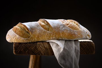 Appetizing sourdough baguette bread with cracked crust and rugged texture on a rustic wooden stool with linen towel. Artisan farmhouse-style loaf captured in warm natural lighting