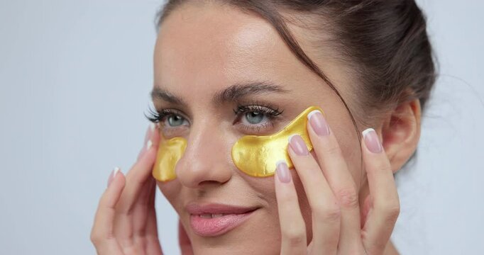 Close up of woman applying gold eye patches sensually posing on camera over gary background. Beauty concept.
