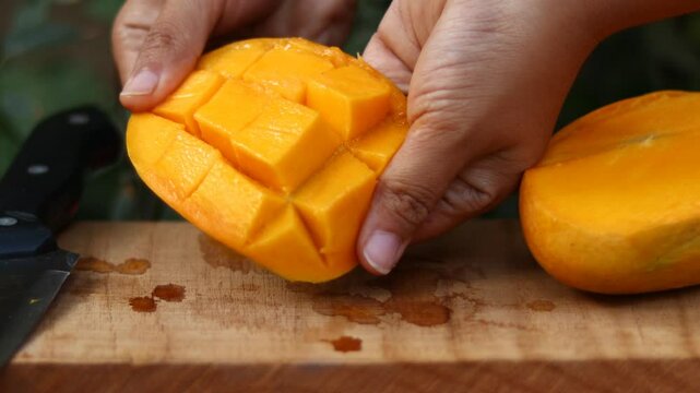 Freshly sliced ripe mango held in hand on wooden cutting board, juicy tropical fruit is cut into cubes - Horizontal clip.