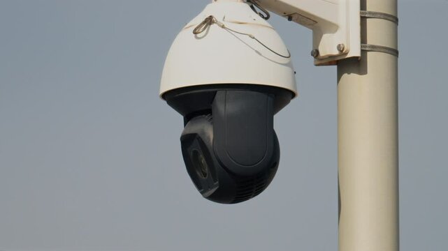 Close up of a round, black and white surveillance camera mounted on a white pole with the cloudy sky on the background