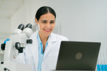 scientist and researcher in laboratory setting smiles while working on laptop, with microscope and...