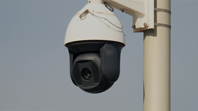 Close up of a round, black and white surveillance camera mounted on a white pole with the cloudy sky on the background