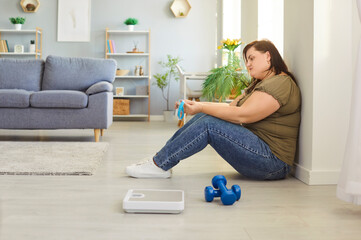 Fototapeta premium Plus-size, fat woman sad as she struggles with fitness and training, sitting on floor at home gym. Emotional challenges of weight loss, workouts, and staying motivated for wellness.