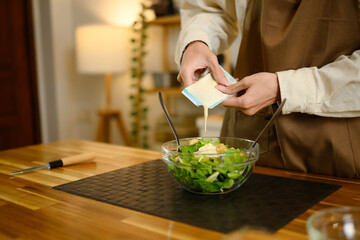 Close up of person pouring salad dressing into a fresh green salad bowl in kitchen