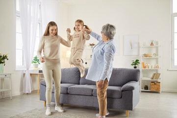 Three female generations have fun together, spend spare time at home. Little girl jumps and holds...