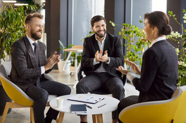 Business meeting in modern office. Two men and woman in suits engaging in productive discussion, smiling and clapping, celebrating successful deal, making agreement, expressing support and approval.