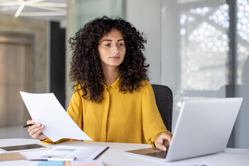 A young woman in a yellow blouse sits at a desk, working on a laptop and reviewing documents. She...