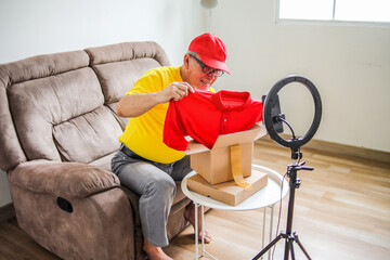 Portrait Of Elderly Male Review Product With Unboxing Package Showing Red Clothes While Doing Vlog