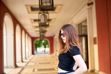Beautiful young woman in black shirt and white skirt standing in front of building with red arch. She holds black handbag and wears sunglasses. Concept of elegance and sophistication. 