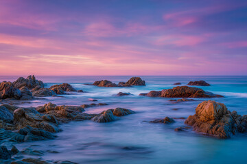 Naklejka premium Long exposure of rocky coastline at sunset with soft pink and blue pastel tones in the sky and sea, evoking a peaceful and dreamlike atmosphere