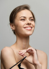 Cheerful young woman with natural makeup, smiling while playfully adjusting her dress strap against a soft gray background. Joy and beauty concept.