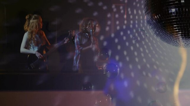Singer leaning into mic while bassist playing bass on moody club stage reflecting disco ball dots