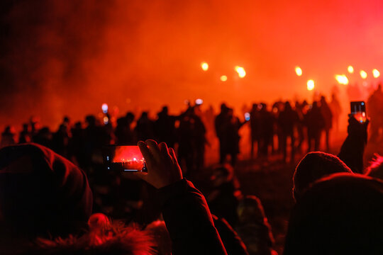 A person holds up a phone to photograph a crowd of people silhouetted against a fiery red sky. Krampus and St Nicholas, 5 December in Camporosso Valcanale, Tarvisio, Italy.